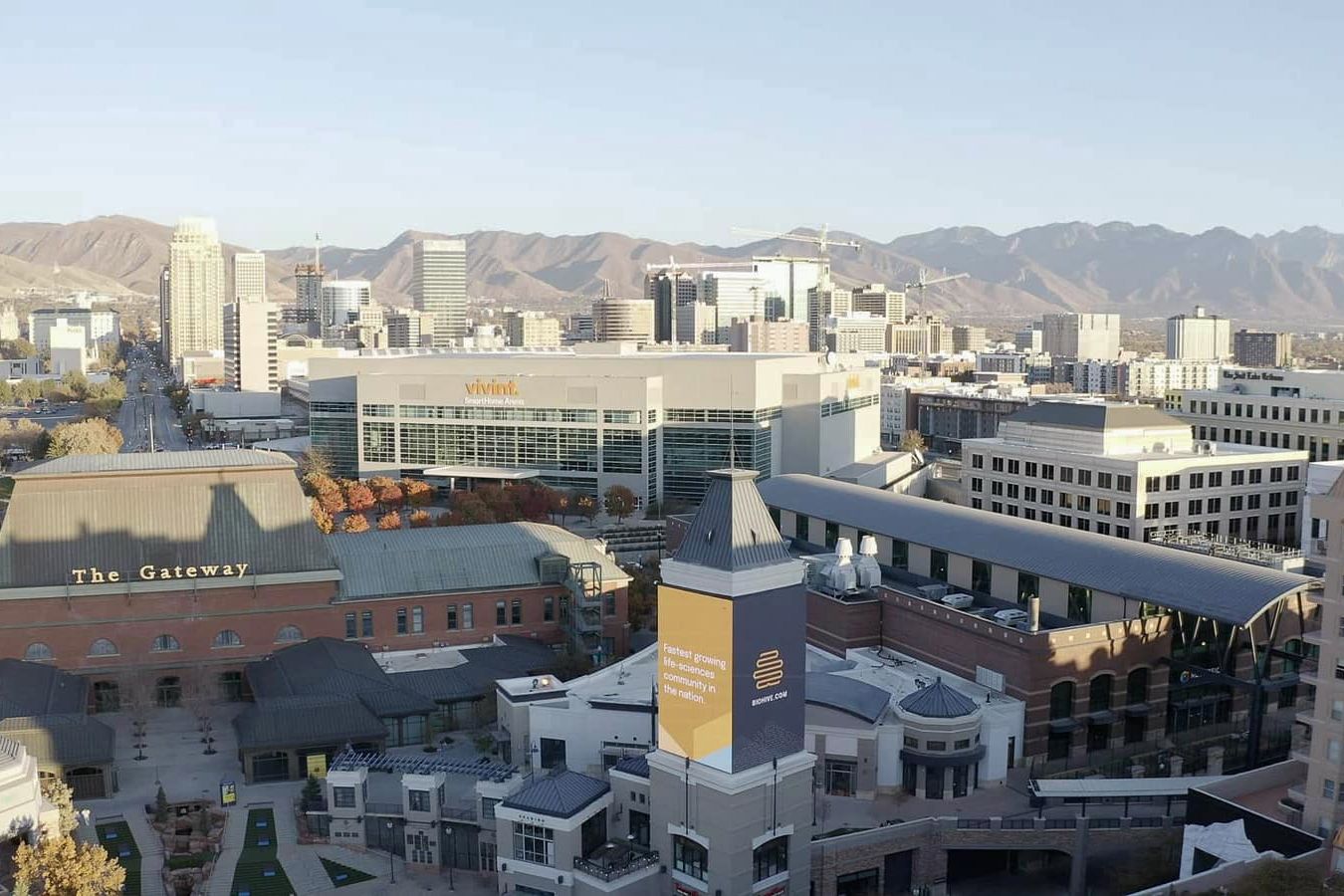 Aerial view of part of Salt Lake City, including the Gateway BioHive complex in the front, skyscrapers behind that, and mountains in the background.