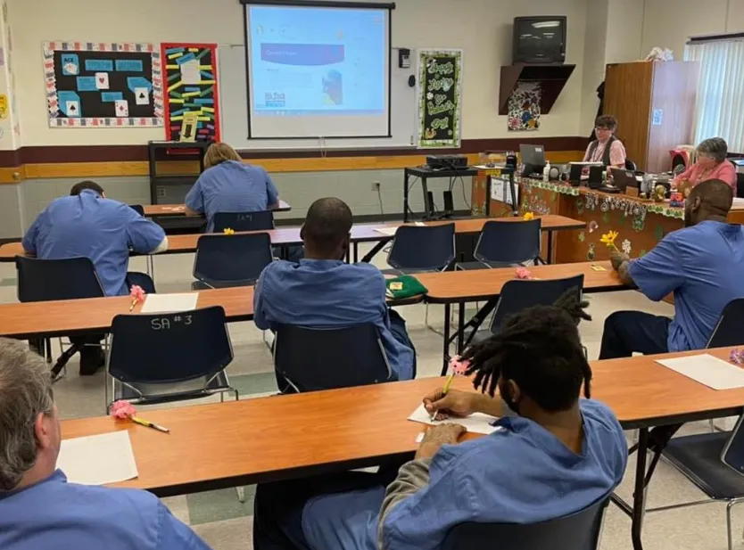 Rear view of several people sitting in desks in a classroom and looking at displays on a TV screen.