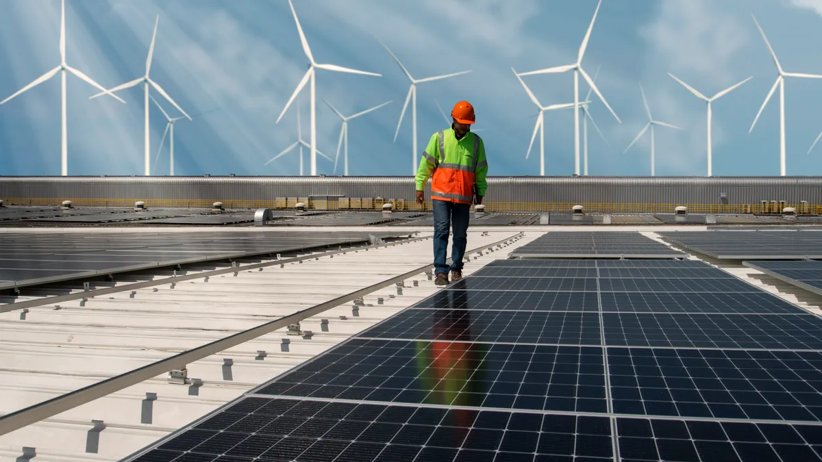 An engineer walks by solar panels with wind turbines in the background.