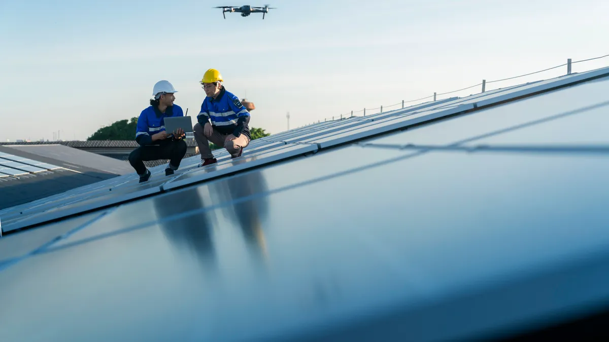 Two technicians work on a solar panel as a drone flies overhead.