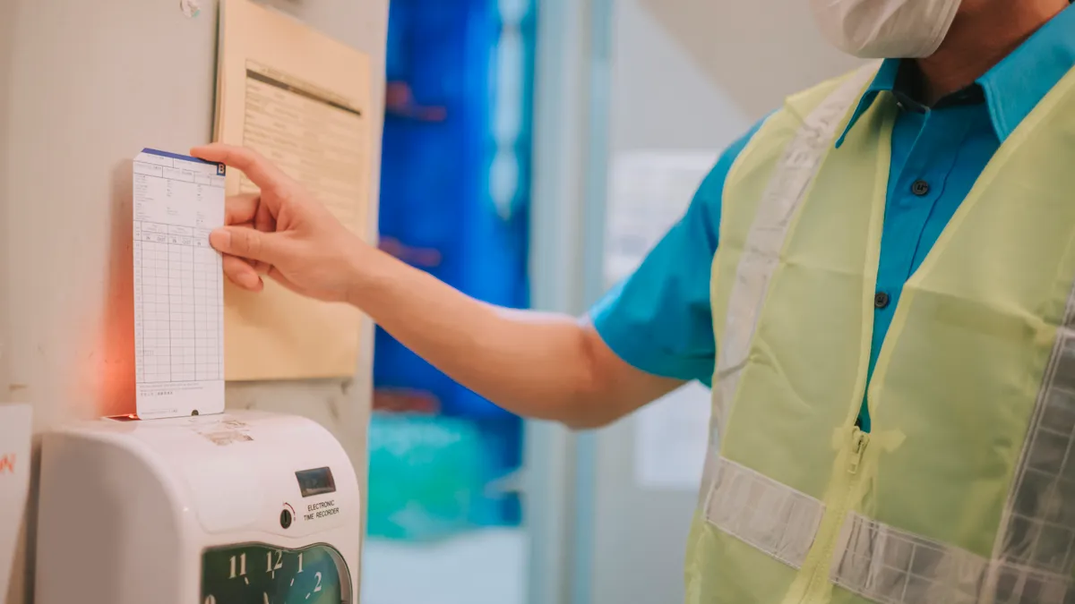 A worker in a safety vests uses a time card machine.