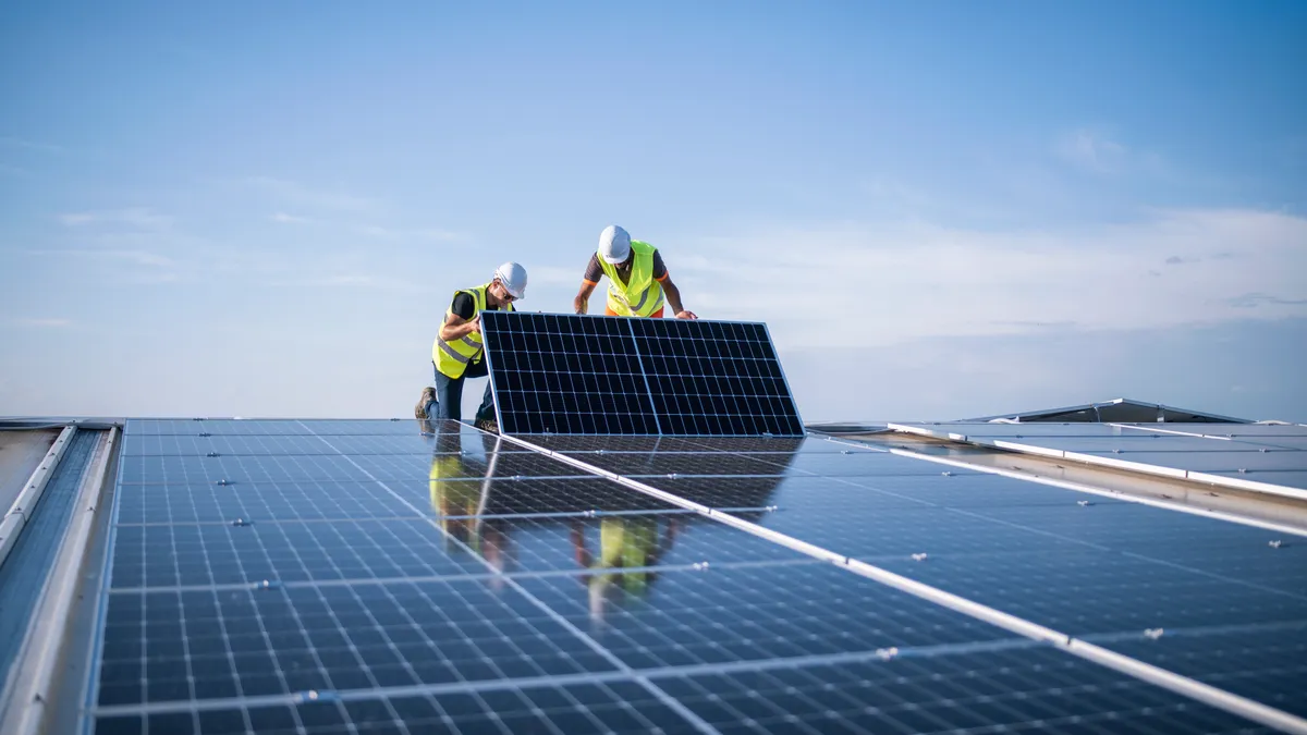 Two workers in hardhats and high-vis vests install a solar panel on a roof.