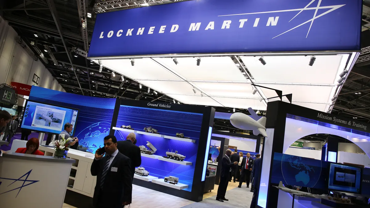 People walk past a Lockheed Martin exhibit in a building.