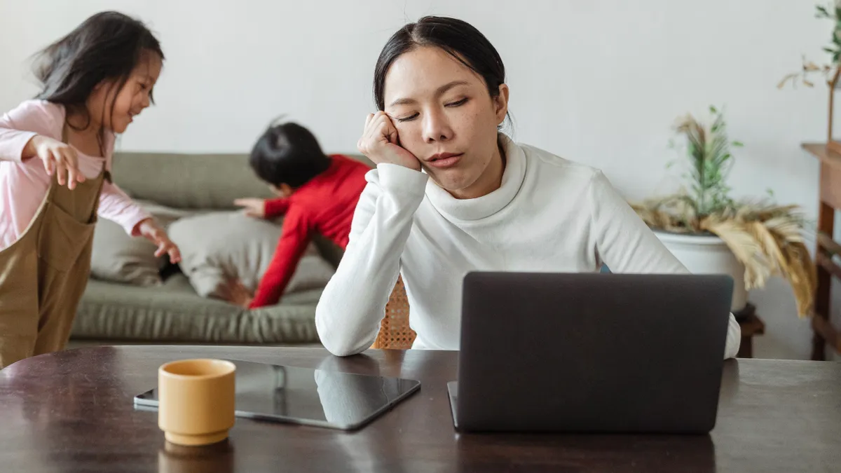 A working parent, surrounded by children, sits tiredly at a laptop